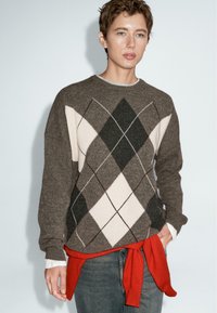 Persona con capelli corti che indossa un maglione argyle marrone, jeans blu e un maglione rosso legato in vita, davanti a uno sfondo semplice.