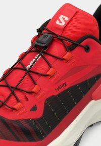 Κόκκινο αθλητικό παπούτσι με επάνω μέρος από διχτυωτό υλικό και μαύρες λεπτομέρειες, σύστημα γρήγορου δέσιμου κορδονιών και μαύρη και κόκκινη σόλα με σχέδιο για καλύτερη πρόσφυση.