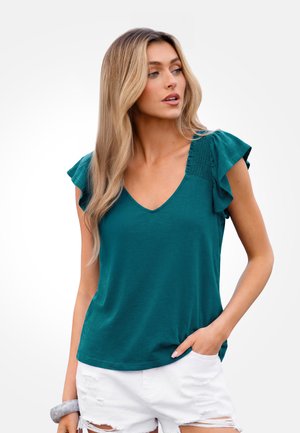Vrouw met lang blond haar die een teal kortemouwtop met gerimpelde schouders en witte gescheurde shorts draagt, kijkt naar de zijkant.