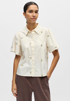 Kvinde iført cremefarvet, kortærmet skjorte med små blomsterbroderier, kombineret med brune bukser, stående med den ene hånd i lommen.