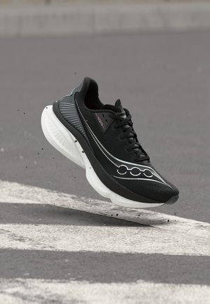 Μαύρο αθλητικό παπούτσι με χοντρή λευκή σόλα, στον αέρα πάνω από ασφάλτινο δρόμο με λευκές γραμμές, μικρά σωματίδια σκόνης να σκορπίζονται από κάτω του.