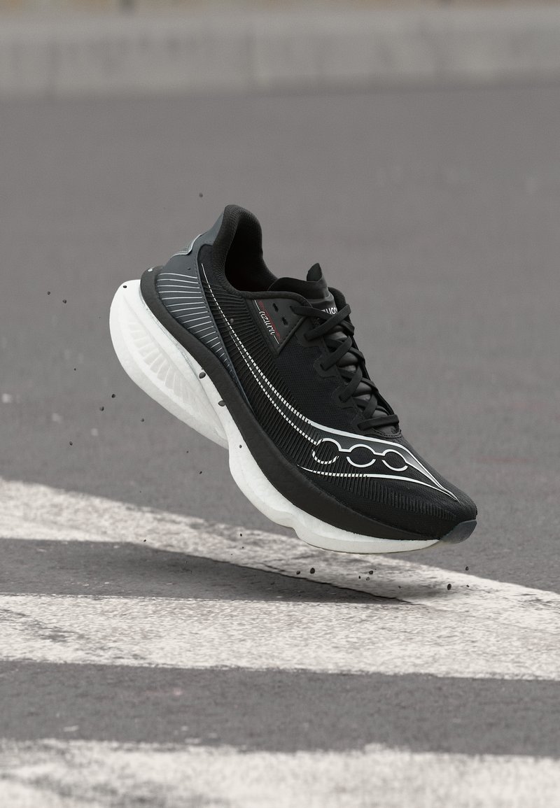 Μαύρο αθλητικό παπούτσι με χοντρή λευκή σόλα, στον αέρα πάνω από ασφάλτινο δρόμο με λευκές γραμμές, μικρά σωματίδια σκόνης να σκορπίζονται από κάτω του.