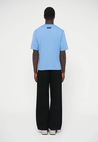 Šviesiai mėlynas medvilninis marškinėlis trumpomis rankovėmis, su mažu juodu etikete prie kaklo, derinamas su juodomis plačiomis kelnėmis ir batais.