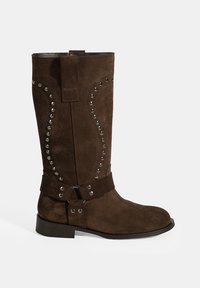 Bottes en suede marron jusqu'au genou avec des clous en métal décoratifs sur les côtés, une large sangle avec des éléments en métal et un design à bout arrondi.