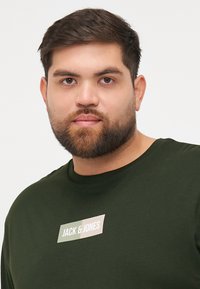 Tamnozelena majica s okruglim izrezom, s svijetlosmešanim pravokutnim logotipom "JACK & JONES" u središnjem dijelu. Mekani teksturirani materijal.
