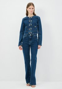 Chaqueta de mezclilla azul oscuro con botones dorados y dos bolsillos frontales, combinada con jeans acampanados a juego que cuentan con acentos de botones en los laterales.