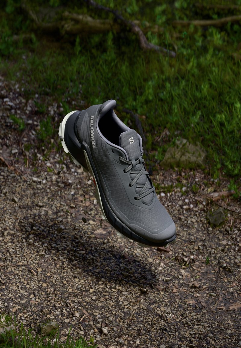Chaussure de course grise avec un dessus texturé, un système de laçage et une semelle rembourrée. Présente le logo de la marque sur le côté et un design léger.