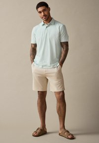 Lys grøn polo-shirt med mønstret tekstur, beige shorts og brune slidesandaler. Casual sommerbeklædning velegnet til varmt vejr.