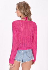 Rosa gehäkelter Pullover mit langen Ärmeln, offenem Strickmuster und einer Krawattenverschluss hinten. Kombiniert mit Denim-Shorts.