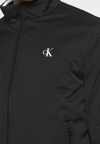 Veste zippée noire en matériau lisse, avec un col montant et un petit logo "CK" blanc sur la poitrine. Deux poches latérales.