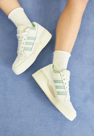 Ben iført hvide platformsneakers med lysegrønne striber og hvide sokker på en blød blå overflade.