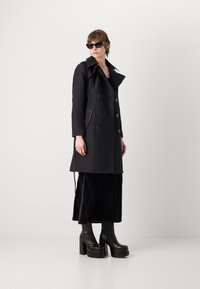 Manteau noir en trench avec des boutons double boutonnage, des poches latérales et un col. Associé à une longue jupe noire et des bottes à talons épais.
