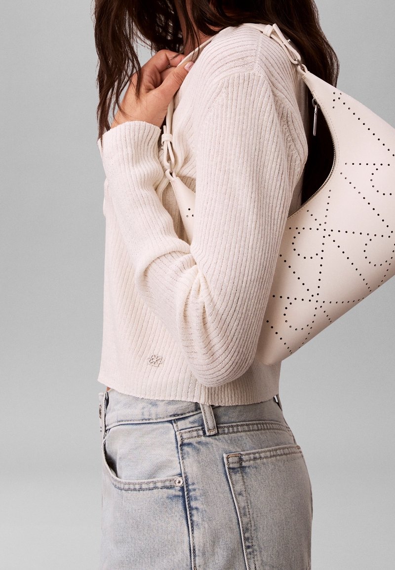 Femme portant un pull côtelé crème et un jean bleu clair, tenant un sac à bandoulière en cuir blanc perforé.