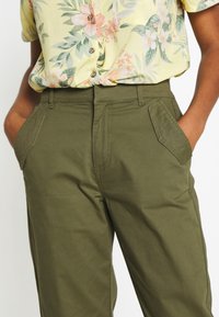 Olivově zelené, středně vysoké kalhoty s předními kapsami a prošívanými detaily. Nošeny s květinovou žlutou košilí s krátkým rukávem. Bavlněný materiál.
