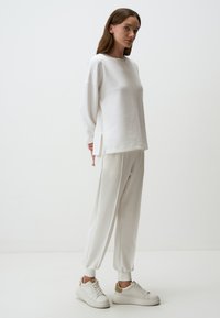 Witte sweatshirt met zijsplitten, gecombineerd met bijpassende witte tapered broek. Schoeisel: lichte sneakers met tan accenten, minimalistisch ontwerp.