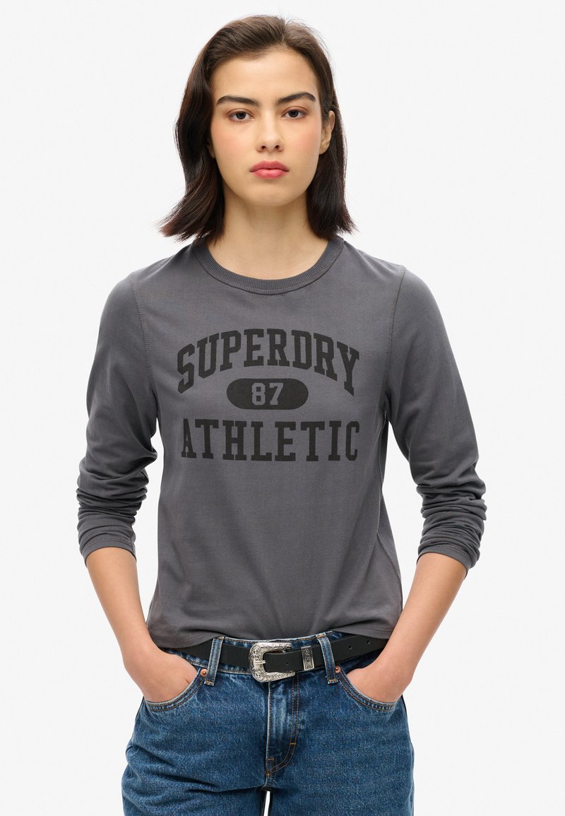 Grå langærmet t-shirt med fed sort "SUPERDRY 87 ATHLETIC" tekst. Båret med højtaljede blå jeans og et sort bælte.