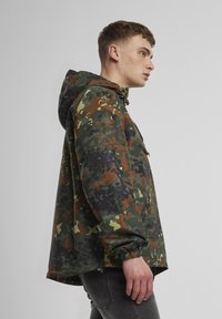 Young man wearing green, brown, and black camouflage hooded jacket with side zipper pocket, standing in profile against a plain background.