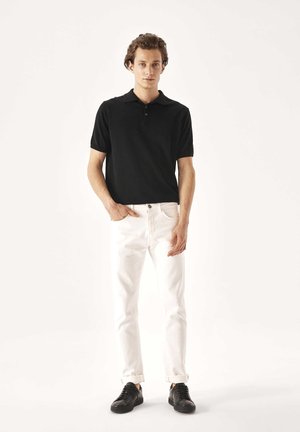 Giovane uomo in piedi, indossa una polo nera a maniche corte, jeans bianchi risvoltati e sneakers nere, mani in tasca su uno sfondo chiaro e uniforme.