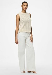 Stickad ärmlös topp i beige med mönster i öppen väv, parat med vita vida byxor. Modellen står framför en enfärgad bakgrund.