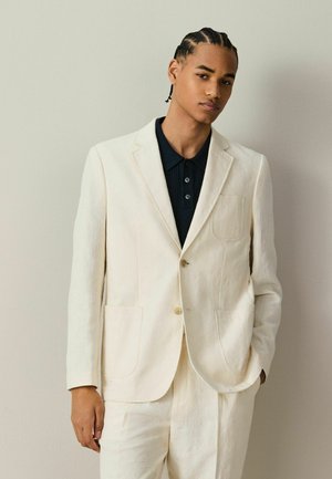 Young man with braided hair wearing a cream suit jacket and pants over a dark collared shirt, standing against a plain light background.