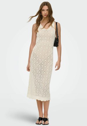 Kvinne iført ermeløs, kremfarget heklet midikjole med diamantmønster, bærer liten svart skulderveske og svarte åpne tå-sandaler.