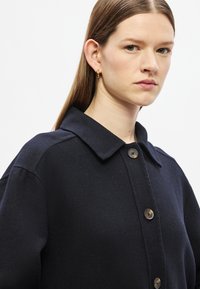 Casaco azul-marinho em mistura de lã com gola de colarinho, botões grandes e corte descontraído. Apresenta textura suave e detalhes subtis de costura.