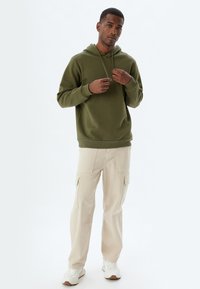 Sudadera verde oliva con cordón, combinada con pantalones cargo beige que tienen bolsillos laterales. El modelo está de pie con zapatillas blancas frente a un fondo claro.