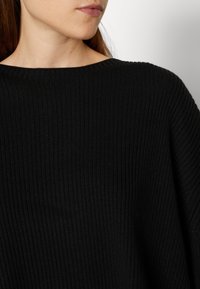 Femme portant un pull en maille côtelée noir avec un large décolleté, montrant une partie de son cou et son visage tourné de côté.