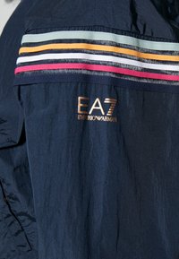 Námořnická bunda má lehkou texturu, zdobenou horizontálními mnohobarevnými pruhy a zlatým logem EA7 na levé straně.