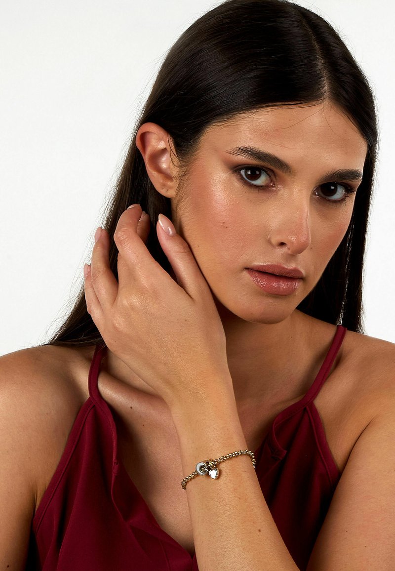 Bracciale dorato realizzato con catena di metallo intrecciata, caratterizzato da charm rotondi e a forma di cuore. Il modello indossa un top senza maniche bordeaux.