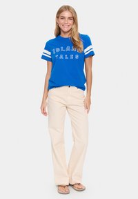 T-shirt en coton bleu avec des rayures blanches et "ISLAND TALES" imprimé en blanc, associé à un pantalon large beige et des tongs brunes.