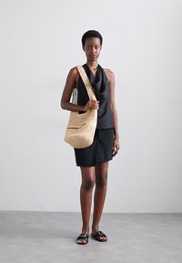 Geantă din paie țesută cu model deschis, de culoare crem, cu curea lungă și numele brandului embosat. Modelul poartă un top negru și o fustă texturată neagră.