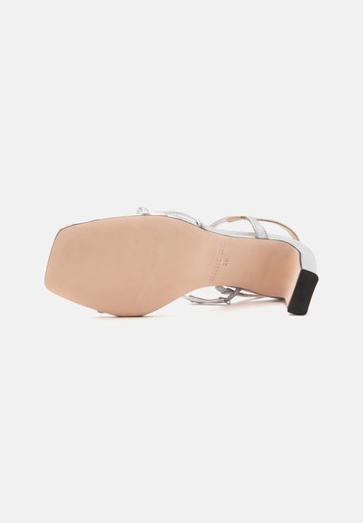 Sandale à bout carré, semelle beige, lanières supérieures argentées et petit talon noir, vue de dessous sur fond blanc.