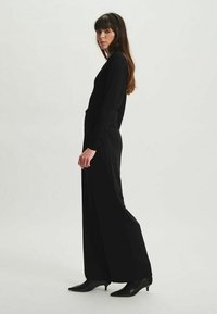 Robe noire à manches longues, comportant un haut ajusté et un bas à jambes larges, fabriquée dans un tissu lisse avec un design minimaliste. Accentuée par des chaussures à talons hauts.