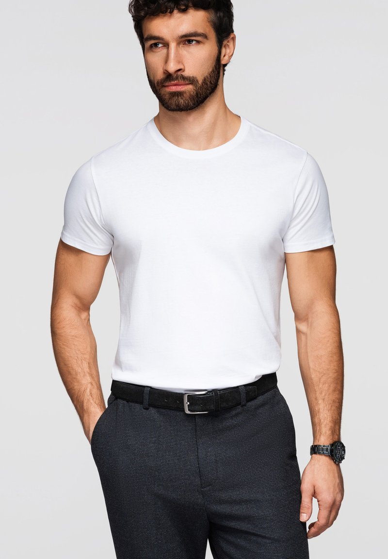 Homme barbu portant un t-shirt blanc uni rentré dans un pantalon foncé, ceinture noire et montre-bracelet noire, debout avec une main dans la poche.