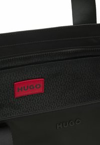 Black bag interior showing a zipper pocket, patterned lining with repeated "HUGO" text, and a red patch labeled "HUGO."