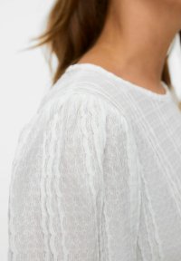 Gros plan sur une personne portant un blouse blanche texturée avec des rayures ondulées verticales et un col rond.