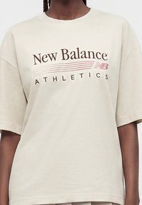 Άτομο που φοράει ένα μπεζ oversized T-shirt με την επιγραφή "New Balance Athletics" και το λογότυπο τυπωμένα στο στήθος σε καφέ και ροζ χρώμα.