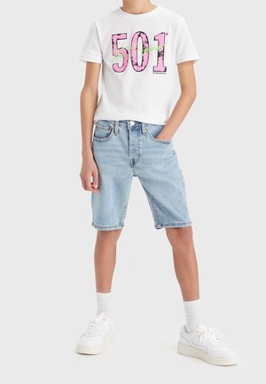Personne portant un t-shirt blanc avec un texte rose « 501 », un short en jean bleu clair, des chaussettes blanches et des baskets blanches, debout avec une main dans la poche.