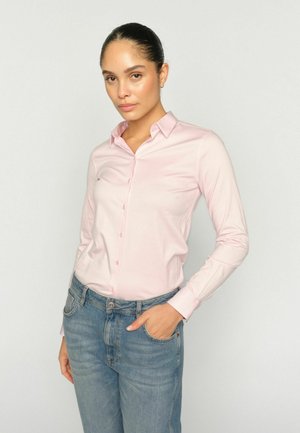 Žena s vlasmi zviazanými dozadu, oblečená v svetloružovej košeli na gombíky a modrých džínsach, stojaca s jednou rukou v vrecku na jednoduchom pozadí.