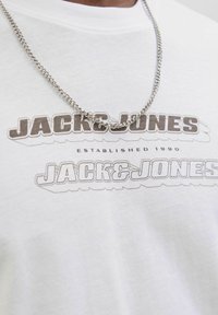 Hvid bomulds T-shirt med "JACK & JONES" logo i brunt, med et trykt mønster. Accentueret med en sølvkæde halskæde.
