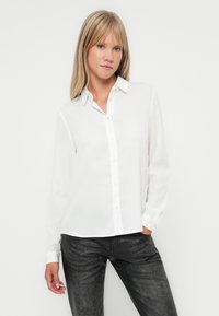 Witte button-up shirt van lichtgewicht stof, met een puntige kraag, lange mouwen en een iets verkorte zoom. Draagt met zwarte gestructureerde broek.