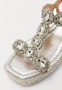 Sandalo metallico argento con upper decorato di gioielli caratterizzato da grandi strass trasparenti, design impreziosito e suola in juta intrecciata.