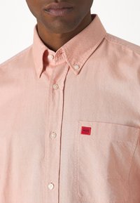 Helles rosa Hemd mit Knopfleiste und einer Brusttasche, weißen Knöpfen und einem roten Logo-Patch. Der Stoff hat eine glatte Textur mit subtilen Webdetails.