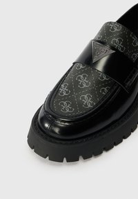 Černé lakované kožené mokasíny s šedým vzorem loga na svršku. Mají robustní podrážku a výrazný kovový akcent loga na pásku.