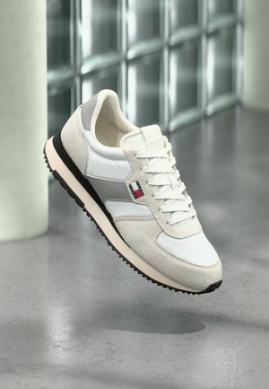 Baskets blanches et grises avec une semelle noire et un petit logo rouge, blanc et bleu, flottant au-dessus d'un sol en béton poli dans un intérieur moderne.