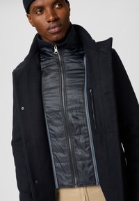 Marinblå ullrock över en marinblå vadderad jacka, med hög krage, dragkedja och ribbade detaljer. Texturerad och strukturerad passform.