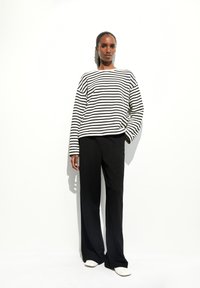 Zwart-wit gestreept shirt met lange mouwen en losse pasvorm, gecombineerd met zwarte wijde broekspijpen en witte instapschoenen. Effen achtergrond.