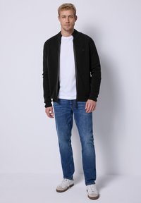 Svart zip-jacka över en vit t-shirt, kombinerad med blå jeans och vita sneakers. Jackan har en rund krage och ribbad mudd.