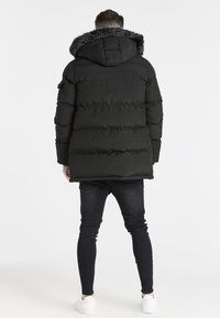 Chaqueta acolchada negra con capucha y ribete de piel sintética, con diseño acolchado y puños acanalados. Combinada con jeans oscuros de corte ajustado y zapatillas blancas.